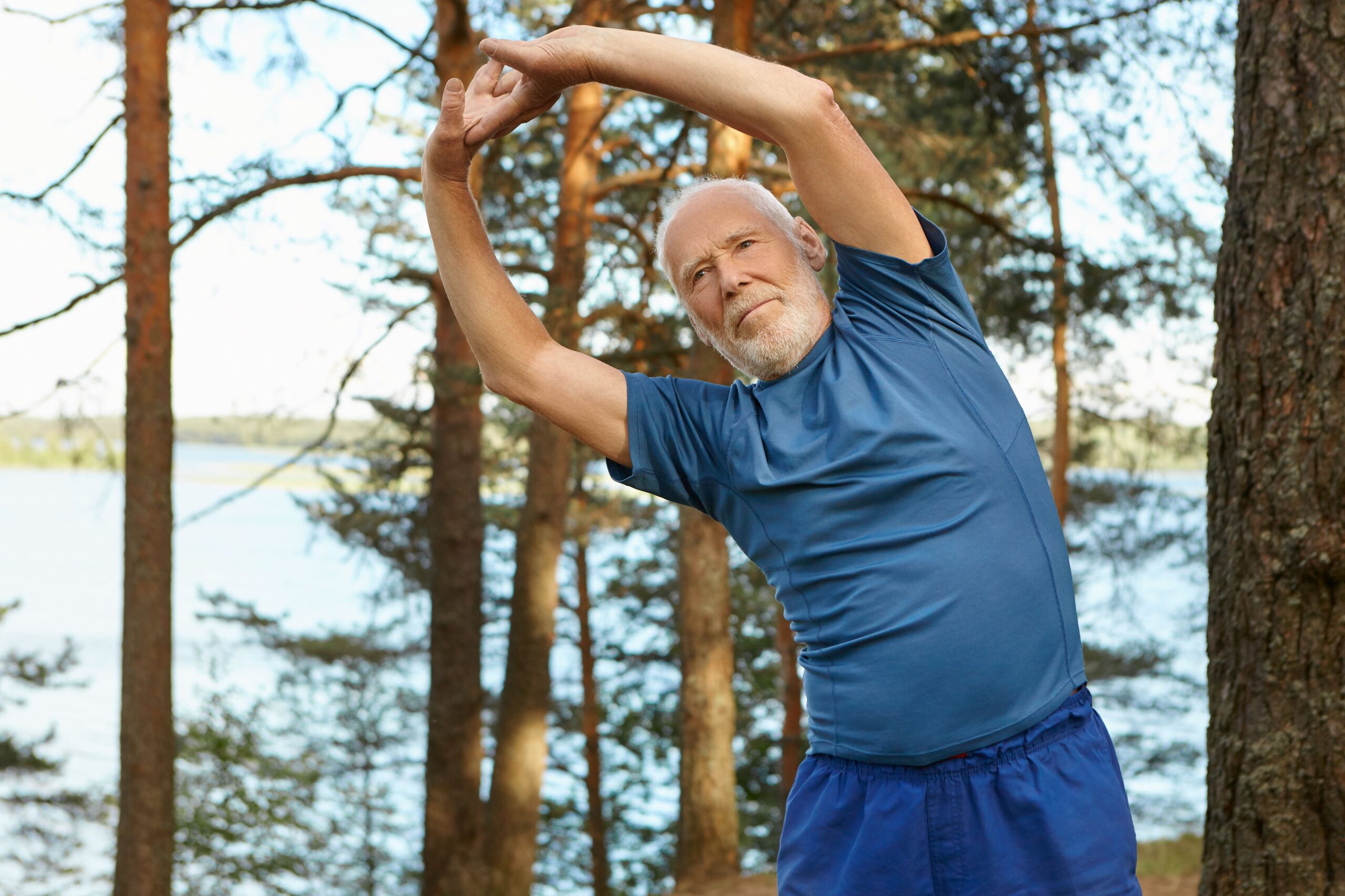 energetic self determined elderly bearded man sportswear posing outdoors with forest river keeping arms raised doing side bends and preparing for senior physical exam